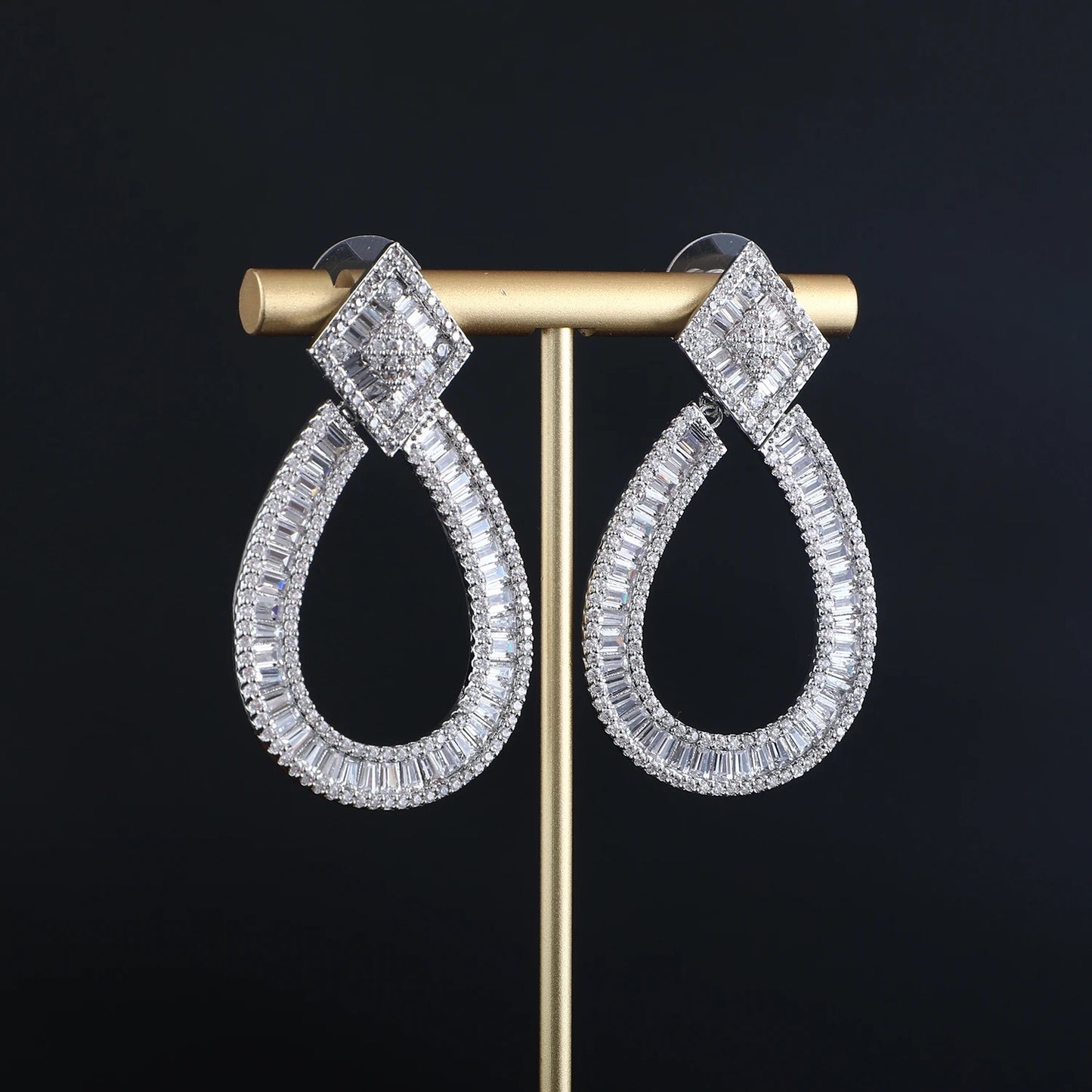 The SONYA EARRINGS by LUCKY13 are a pair of elegant teardrop-shaped drop earrings encrusted with clear, shimmering crystals. Each earring has a geometric, square diamond-like design at the top, connected to a wider teardrop shape adorned with sparkling rectangular stones. These statement pieces hang on a gold stand.