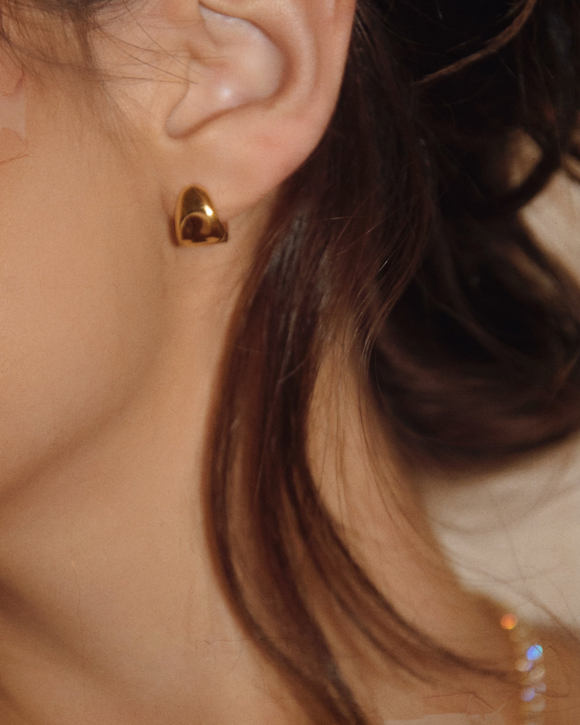 Close-up of a person’s ear adorned with a small, smooth, lightweight gold earring. The earring has a teardrop shape and the person's hair is dark and slightly wavy, partially covering the ear. The lighting is warm, highlighting the comfortable PUFFY HUGGIES by LUCKY13 and hair.