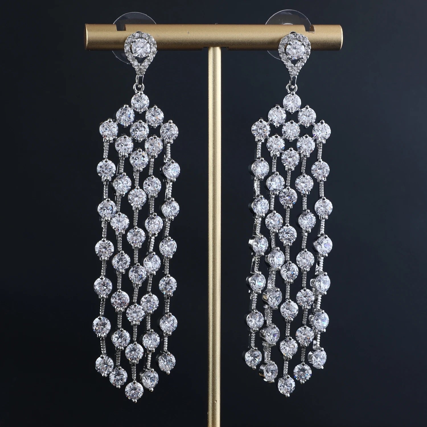 The NIAGARA EARRINGS by LUCKY13 are a pair of luxurious, dangling earrings adorned with multiple rows of sparkling cubic zirconia gemstones, each row connected by smaller stones. The top features a circular stud design in hypoallergenic sterling silver, and the earrings are displayed on a golden stand against a black background.