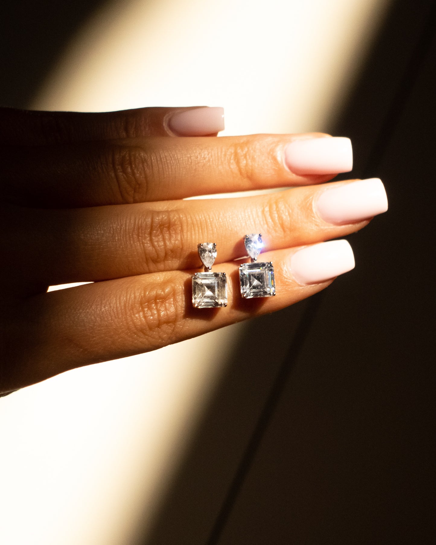 A woman hand holding a pair of elegant drop earrings featuring pear-shaped and square-cut gemstones, crafted from 925 sterling silver.