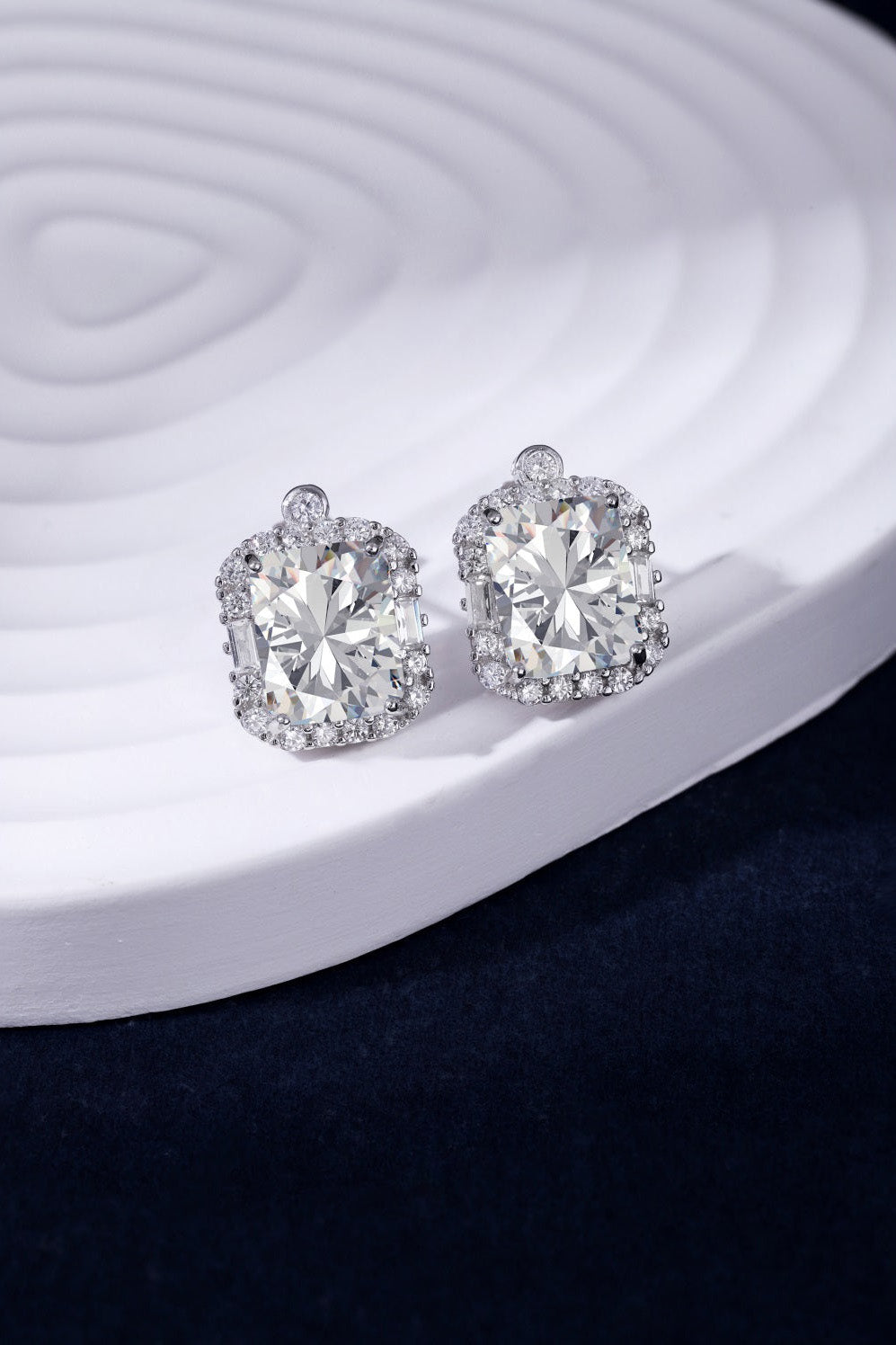 A pair of elegant VINTAGE DIAMOND STUDS by LUCKY13 with large, radiant-cut cubic zirconia gemstones, each surrounded by a halo of smaller sparkling stones, set on a white, sculpted circular surface against a dark blue background. Hand-finished in 925 sterling silver for an added touch of sophistication.