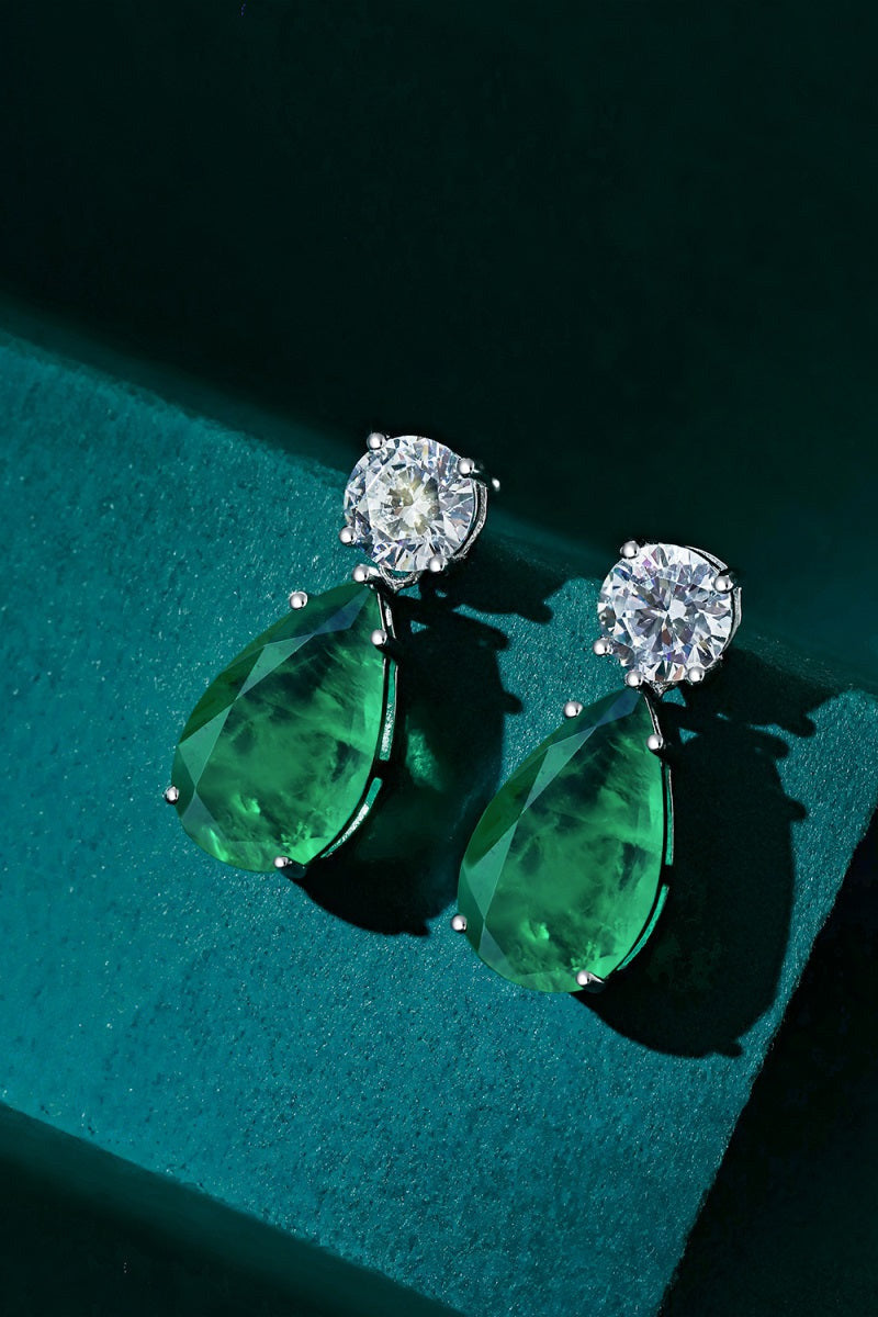 A pair of luxurious, elegant JADE EARRINGS by LUCKY13 featuring large, teardrop-shaped green gemstones suspended below round, clear-cut diamonds, all set in 925 sterling silver. The earrings are showcased against a textured teal background, with shadows highlighting their facets and metallic settings.