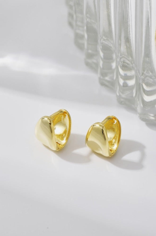 A pair of shiny, gold-toned PUFFY HUGGIES by LUCKY13 rests on a flat, light-colored surface. The lightweight earrings have a smooth, curved design perfect for everyday wear. In the background, there is a transparent, ridged glass object partially visible, adding a reflective element to the scene.