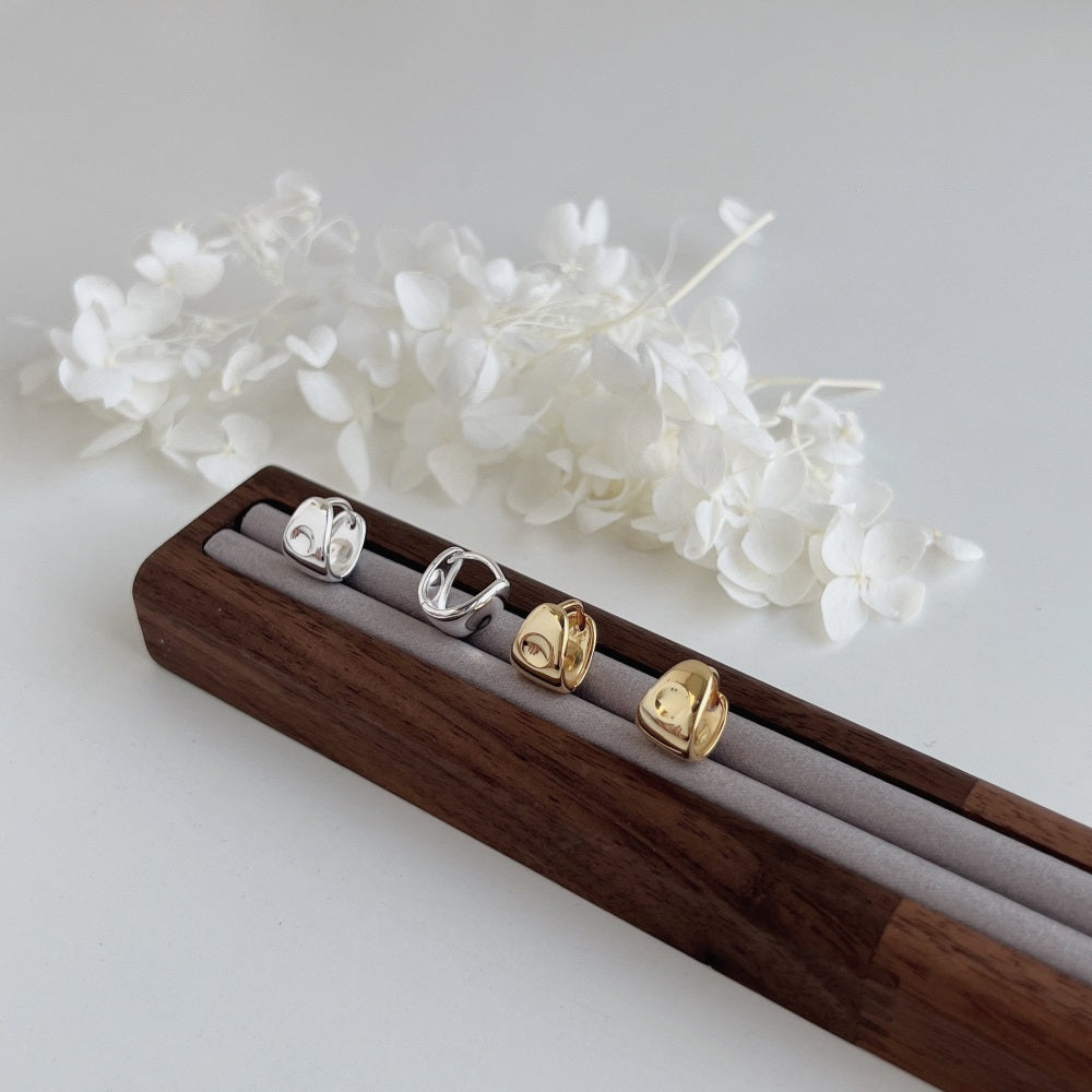 Four small, minimalist rings from the PUFFY HUGGIES collection by LUCKY13—two 14K gold plated and two 925 sterling silver—are displayed on a wooden ring holder with a soft gray fabric insert. In the background, delicate white flowers rest on the smooth white surface.