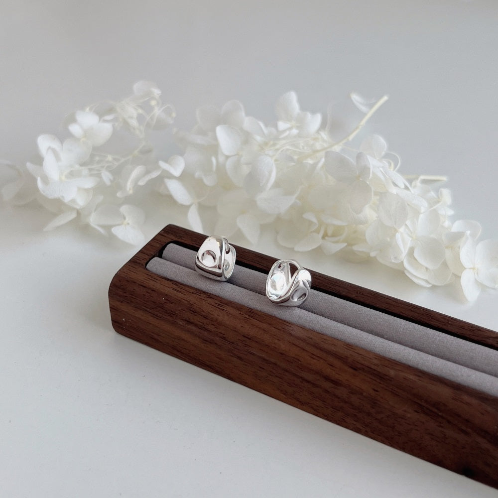 A pair of PUFFY HUGGIES from LUCKY13, featuring 925 Sterling Silver teardrop shapes, rest on a wooden jewelry holder with a gray padded strip. The holder is placed on a white surface, and delicate white flowers are arranged in the background, creating a minimalist and elegant display.